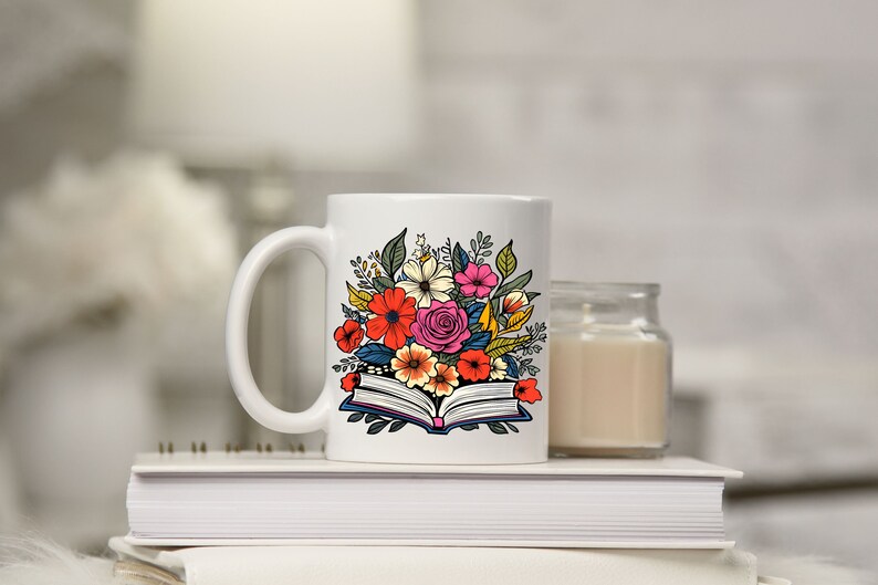 K&ouml;nnte beinhalten: Wei&szlig;e Keramiktasse mit einem bunten Blumenmuster auf einem offenen Buch. Die Tasse steht auf einem Stapel B&uuml;cher.