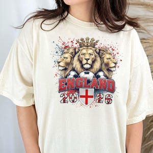 Puede incluir: Camiseta color crema con un gr&aacute;fico de tres leones coronados, balones de f&uacute;tbol y la palabra "ENGLAND" en rojo. Debajo, el a&ntilde;o "2026" y la bandera inglesa. La camiseta tiene un corte holgado.