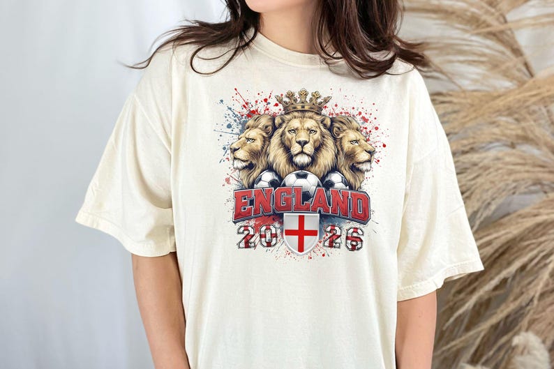 Puede incluir: Camiseta color crema con un gr&aacute;fico de tres leones coronados, balones de f&uacute;tbol y la palabra "ENGLAND" en rojo. Debajo, el a&ntilde;o "2026" y la bandera inglesa. La camiseta tiene un corte holgado.