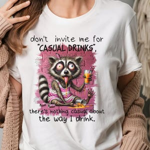 May include: White t-shirt with a cartoon raccoon drinking from a can and the text "don't invite me for "CASUAL DRINKS", there's nothing casual about the way I drink."