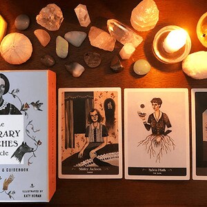 Puede incluir: Una caja azul con el texto "The Literary Witches Oracle" y una baraja de 70 cartas y un libro de instrucciones. La caja está rodeada de cuatro cartas con ilustraciones de mujeres y el texto "Shirley Jackson", "Sylvia Plath" y "Toni Morrison".