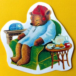 Puede incluir: Una pegatina troquelada con la ilustración de un oso pardo con gorro de dormir rojo y camisón azul claro, durmiendo en un sillón verde. Junto al oso hay una pequeña mesa de madera con una tetera blanca, una taza de té y pasteles.