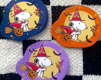 May include: Three stacks of die-cut stickers featuring Snoopy as a witch wearing a purple hat with yellow stars and a pink cape, riding a broomstick with a jack-o'-lantern. The background colours are dark blue, orange, and purple with bats.