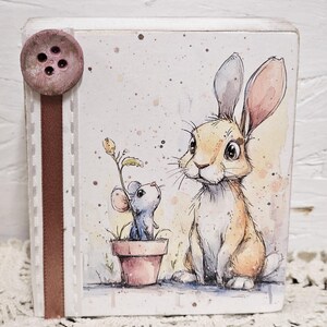 May include: A square wooden block featuring a watercolor illustration of a tan rabbit sitting next to a small blue mouse inside a terracotta pot. A brown ribbon with a circular button is attached to the left side of the white block.