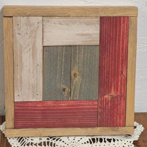 May include: A square wooden wall art piece with a natural wood frame. The interior is composed of various rectangular wooden planks painted in white, red, and gray-green hues. The piece is displayed on a white lace doily.