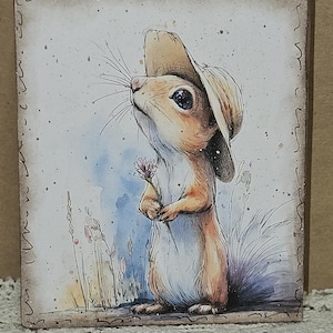 May include: A rectangular wooden block featuring a watercolor illustration of a small squirrel wearing a tan hat and holding a tiny purple flower. The squirrel has large dark eyes and long whiskers, set against a light background with blue accents.