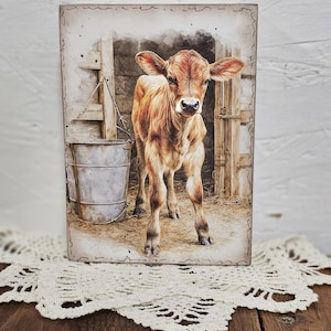 May include: A rectangular artwork depicts a brown calf standing in a barn. The calf has a light brown coat and is looking directly at the viewer. A metal bucket hangs on the left, and a wooden gate is in the background. The artwork is framed with a decorative border.