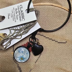 May include: A close-up of cross-stitch supplies, including a black embroidery hoop, a tag of Classic Colorworks Belle Soie thread in Moss, a pendant with a blue floral design, and a black heart-shaped charm. A needle is also visible.