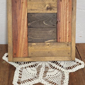 May include: Square wooden frame with a rustic, reclaimed wood design. The frame features a mix of natural wood tones, including reddish-brown, light brown, and dark brown. The frame is sitting on a white crocheted doily.
