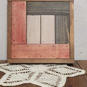 May include: Square wooden wall art with a rustic design. The artwork features a frame and a patchwork of wooden planks in shades of brown, red, gray, and cream. The piece is displayed on a wooden surface.