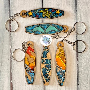 May include: Set of six wooden surfboard keyrings featuring various designs. The designs include a turtle, dolphins, palm trees, and waves. The keyrings are painted in bright colours, including blue, green, yellow, orange, and red.