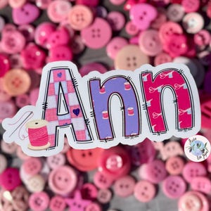 May include: A sticker featuring the name "Ann" in a playful style. The letter "A" is pink and white chequered with a heart, the "n" is purple with thread spools, and the second "n" is pink with scissors. A pink thread spool and needle are on the left.