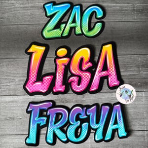 May include: Three colourful, stylised names, "Zac", "Lisa", and "Freya", are written in a rainbow gradient with black outlines and white polka dots. The names are on a wooden background.