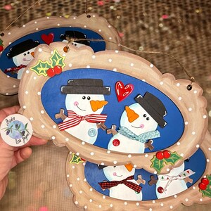 May include: Two wooden snowman ornaments with blue backgrounds and white polka dots. The snowmen are wearing black hats, red and white striped scarves, and blue scarves. They are holding hands and there is a red heart between them. The ornaments are hanging from a string.