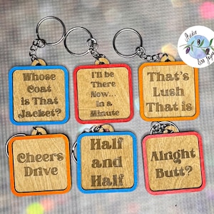 May include: Six wooden keychains with different sayings. The keychains are square and have a light brown wood background with orange, red, and blue borders. The sayings on the keychains are: "Whose Coat is That Jacket?", "I'll be There Now... In a Minute", "That's Lush That is", "Cheers Drive", "Half and Half", and "Alright Butt?"