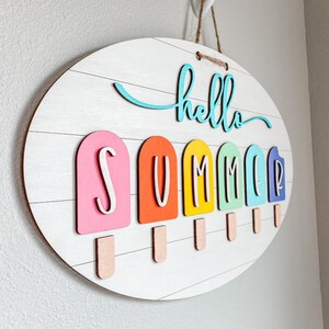 May include: A white oval wooden sign with the words "hello summer" in turquoise blue lettering. The sign has six colorful popsicle-shaped letters spelling out "SUMMER" with a stick attached to each letter.