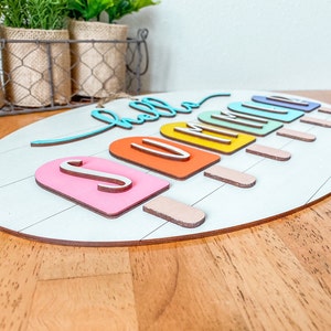 May include: A wooden oval sign with the words "hello summer" in colorful script. The letters are attached to popsicle-shaped pieces of wood in various colors.
