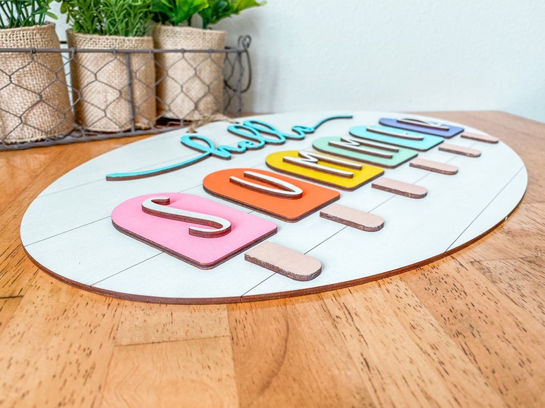 May include: A wooden oval sign with the words "hello summer" in colorful script. The letters are attached to popsicle-shaped pieces of wood in various colors.