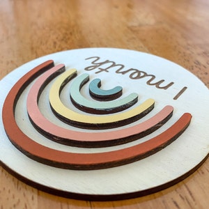May include: A white wooden circle with a rainbow design made of layered painted wood arcs. The text "1 month!" is written in brown cursive on the circle.