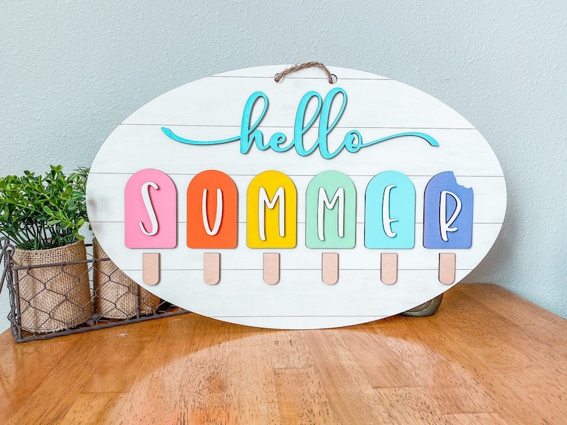 May include: A white oval wooden sign with the words "hello summer" written in blue and white. The word "summer" is spelled out with popsicle shapes in pink, orange, yellow, green, blue, and purple.