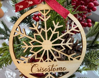 Op de afbeelding: Een houten kerstversiering met een sneeuwvlokdesign en de naam "Crisostomo" erop gegraveerd. De versiering is versierd met een rood lint en omringd door groene dennentakken.