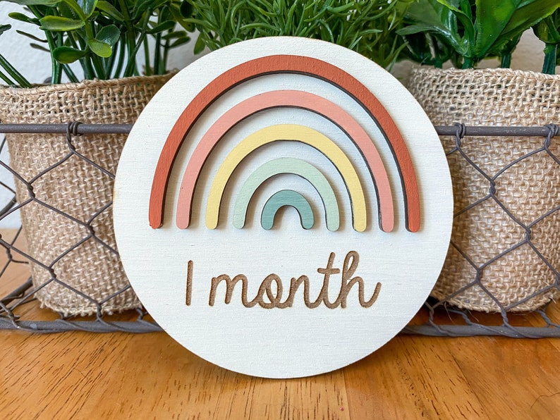 May include: A round wooden baby milestone marker with a rainbow design. The rainbow is made of layered wood pieces in shades of red, pink, yellow, and green. The text "1 month" is engraved below the rainbow.