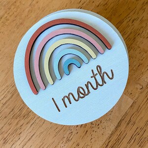 May include: A light blue wooden circle with a rainbow made of different colored wooden pieces and the text "1 month" in brown.
