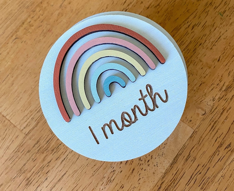 May include: A light blue wooden circle with a rainbow made of different colored wooden pieces and the text "1 month" in brown.