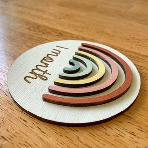 May include: A round wooden ornament with a rainbow design and the text "I am now!" in cursive.