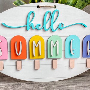 May include: A white wooden sign with the words "hello summer" in turquoise blue lettering. The word "summer" is spelled out with six popsicle-shaped pieces of wood in pink, orange, yellow, green, blue, and purple.