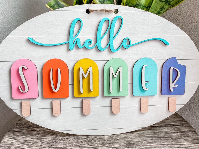May include: A white wooden sign with the words "hello summer" in turquoise blue lettering. The word "summer" is spelled out with six popsicle-shaped pieces of wood in pink, orange, yellow, green, blue, and purple.