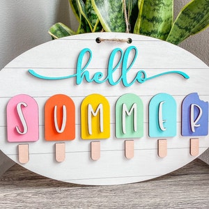 May include: A white oval wooden sign with the text "hello summer" in turquoise lettering. The word "summer" is spelled out with six colorful popsicle-shaped pieces.