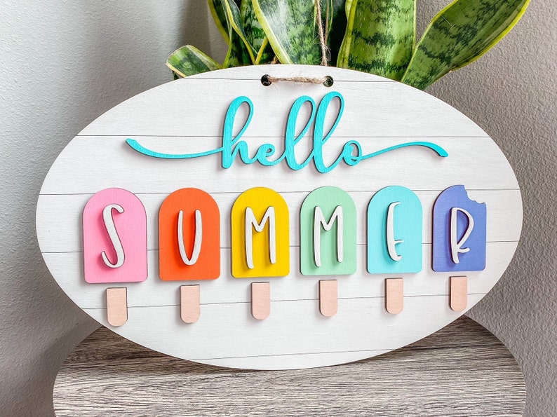 May include: A white oval wooden sign with the text "hello summer" in turquoise lettering. The word "summer" is spelled out with six colorful popsicle-shaped pieces.