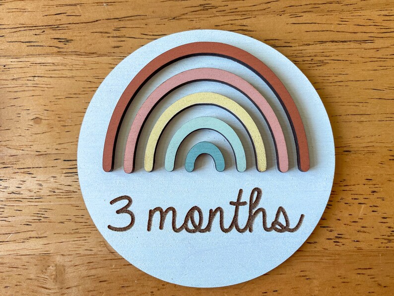 May include: A light blue wooden circle with a rainbow made of layered wooden arcs in shades of brown, pink, yellow, and blue. The text "3 months" is engraved below the rainbow.