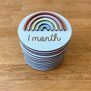 May include: A stack of white wooden circles with a rainbow design and the text "1 month" on the top circle.
