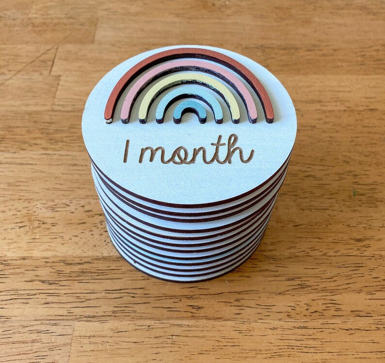 May include: A stack of white wooden circles with a rainbow design and the text "1 month" on the top circle.