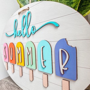 May include: A round wooden sign with the words "hello summer" written in turquoise blue. The word "summer" is spelled out with individual letters on popsicle shapes in various colors: orange, yellow, green, blue, and purple.