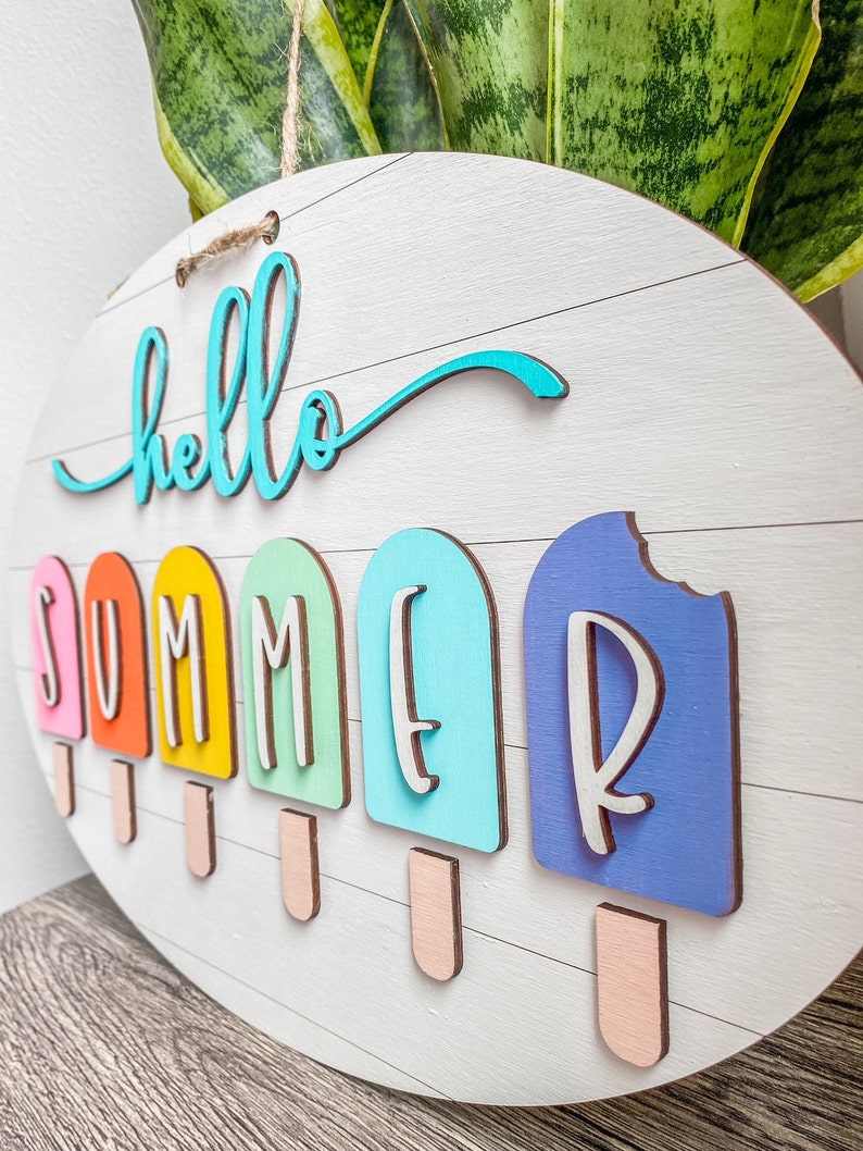 May include: A round wooden sign with the words "hello summer" written in turquoise blue. The word "summer" is spelled out with individual letters on popsicle shapes in various colors: orange, yellow, green, blue, and purple.
