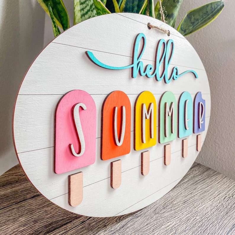 May include: A white wooden oval sign with the words "hello summer" in blue script. The word "summer" is spelled out with six colorful popsicle-shaped pieces.