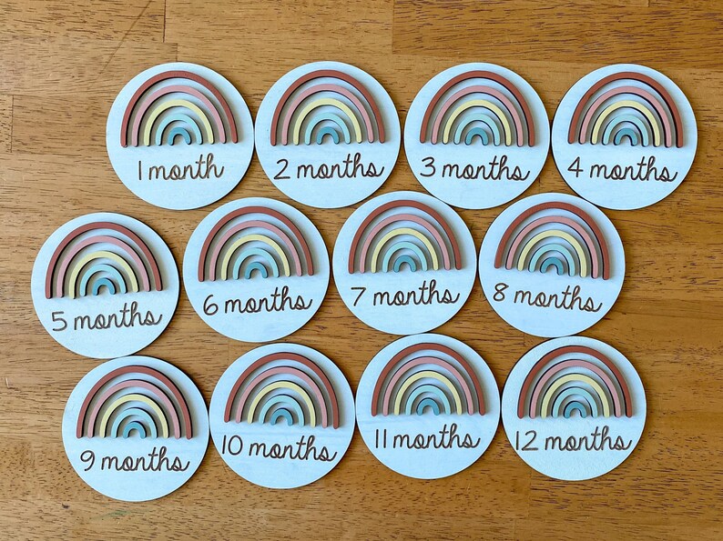 May include: Twelve wooden rainbow-themed baby milestone discs with white backgrounds. Each disc features a rainbow with different colored stripes and a number from 1 to 12, indicating the month of the baby's age.