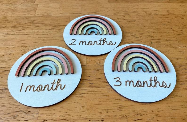 May include: Three round wooden baby milestone markers with rainbow designs. Each marker features a different number of months: 1 month, 2 months, and 3 months.