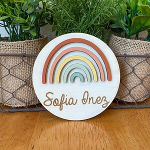 May include: A round wooden sign with a rainbow design in shades of red, orange, yellow, green, and blue. The sign has the name "Sofia Inez" written in cursive.