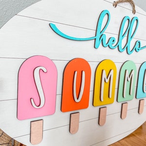 May include: A white wooden sign with the words "hello summer" in blue and white lettering. The word "summer" is spelled out with colorful popsicle-shaped letters.