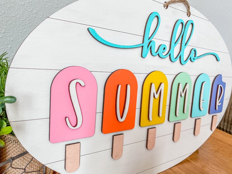 May include: A white wooden sign with the words "hello summer" in blue and white lettering. The word "summer" is spelled out with colorful popsicle-shaped letters.