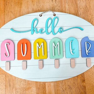 May include: A wooden oval sign with the words "hello summer" written in blue. The word "summer" is spelled out with individual letters on popsicle sticks in various colors: pink, orange, yellow, green, blue, and purple.