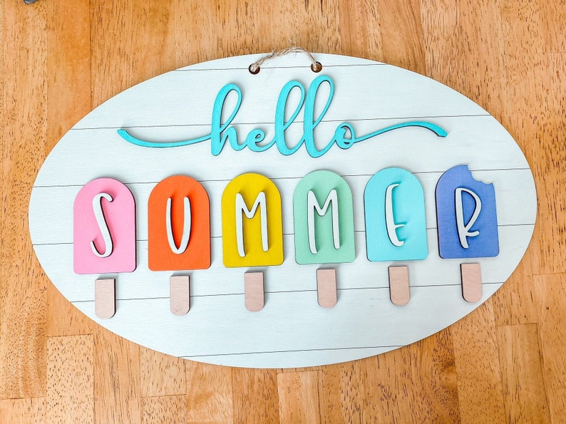 May include: A wooden oval sign with the words "hello summer" written in blue. The word "summer" is spelled out with individual letters on popsicle sticks in various colors: pink, orange, yellow, green, blue, and purple.