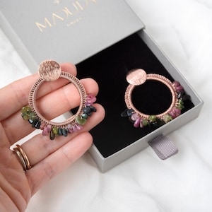 May include: Rose gold hoop earrings with a cluster of pink, green, and black gemstones hanging from the bottom.