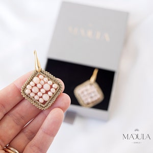 Può includere: Un paio di orecchini dorati con un design quadrato con perline rosa. Gli orecchini sono esposti su una mano e in una scatola con la scritta "MADULA" stampata su di essa.