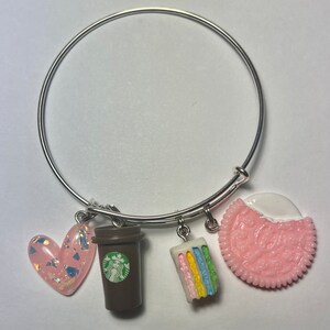 Può includere: Un braccialetto rigido argentato con quattro ciondoli: un cuore rosa con glitter dorati, una tazza di caffè marrone con un logo verde, una torta a strati arcobaleno e un biscotto rosa e bianco.