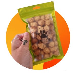 May include: A clear plastic bag of dog treats with a paw print logo and the text "DOGRAZE". The bag is held against a yellow and orange background. The treats are small and round, and the bag has a green top.